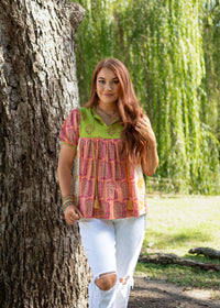 Woman with red hair standing front profile in lifestyle scene that has heavy greenery in Jeanie Top. She is wearing the Jeanie Top paired with white pants. 