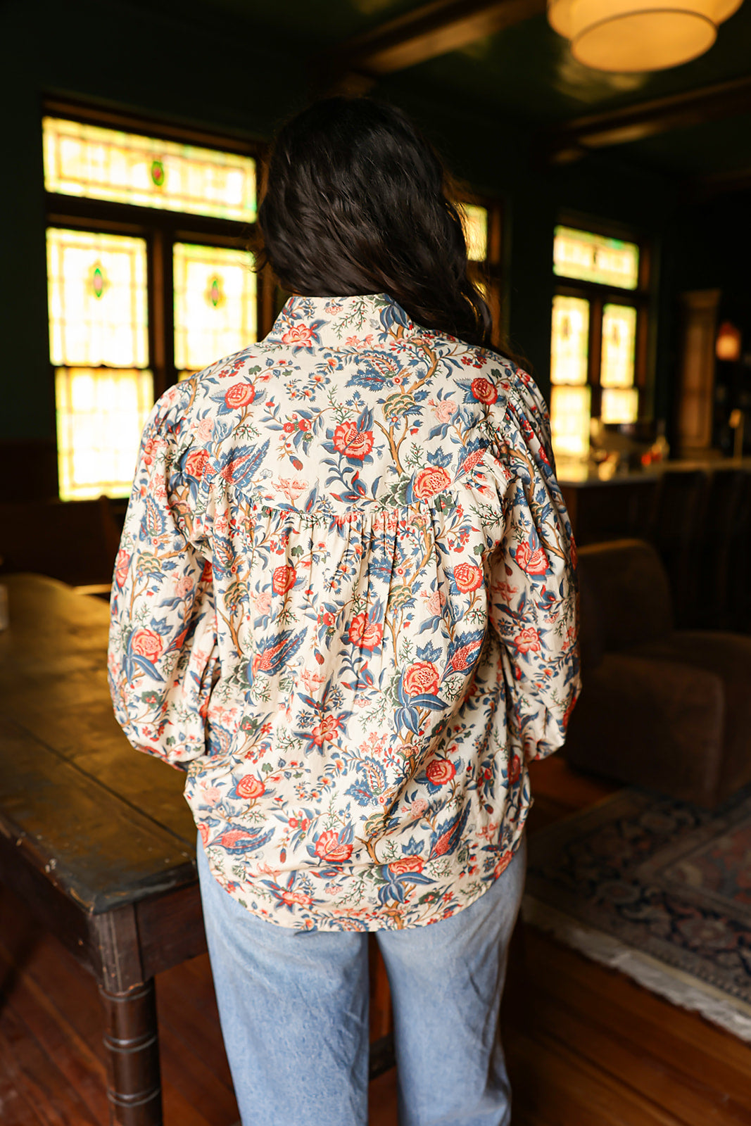 Woman with dark hair standing in a back profile position wearing the Jenna Top in a lifestyle scene. The focal point of this image is the red and blue floral pattern. The model paired this top with light-wash denim jeans. 