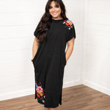 Woman with dark hair standing in a front profile position wearing the Jo Dress against a white background. The focal point of this image is the embroidery on sleeve and bottom of dress. The model paired this dress with white and gold sneakers.