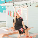 Woman with blonde hair sitting in a front facing position in a lifestyle scene wearing the Jo Dress. The focal point of this image is the front of the dress with slight emphasis on the background that expresses bold colors. The model paired this dress with colorful floral sandals.
