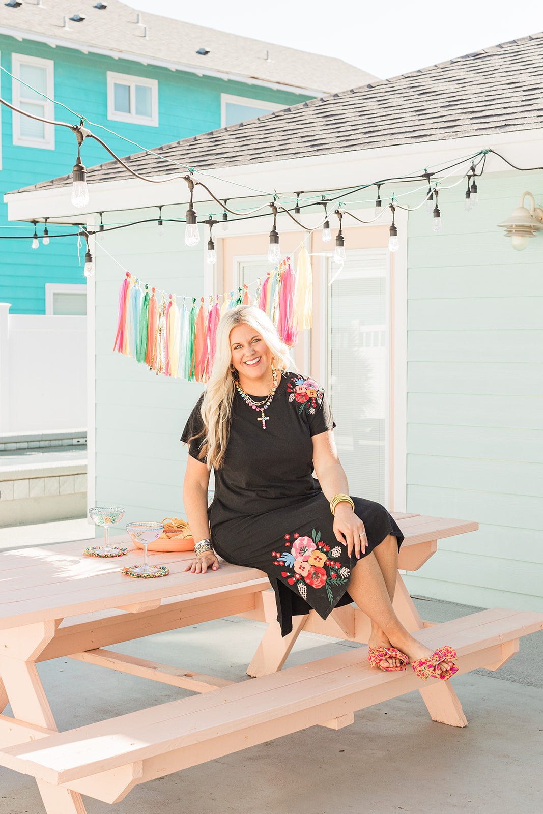 Woman with blonde hair sitting in a front facing position in a lifestyle scene wearing the Jo Dress. The focal point of this image is the front of the dress with slight emphasis on the background that expresses bold colors. The model paired this dress with colorful floral sandals.