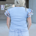 Woman with blonde hair wearing denim top with burnt orange embroidery on sleeves, and light-wash denim shorts.