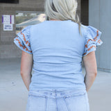 Woman with blonde hair wearing denim top with burnt orange embroidery on sleeves, and light-wash denim shorts.
