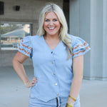 Woman with blonde hair wearing a denim top with burnt orange embroidery on sleeves, and light-wash denim shorts.