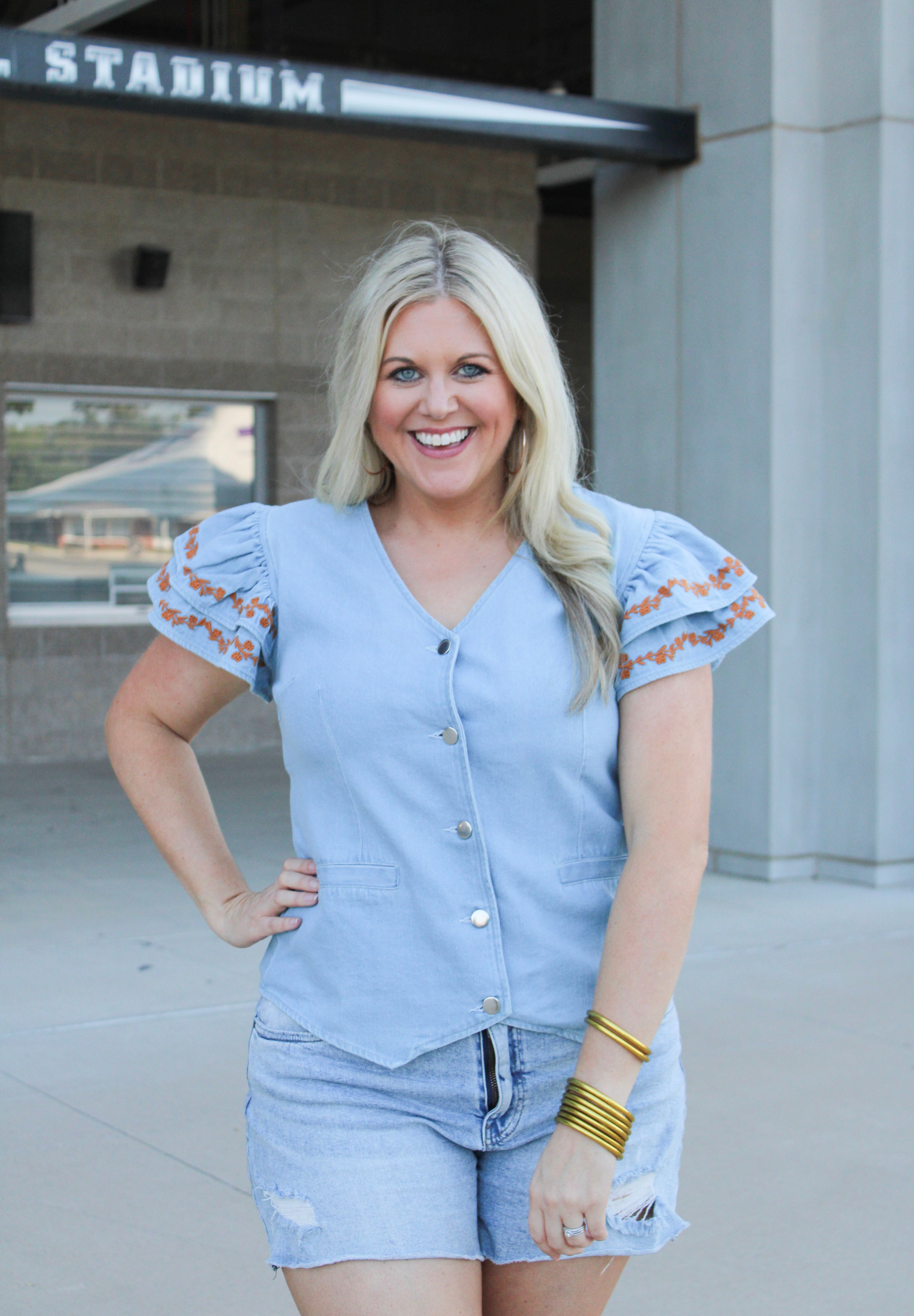 Woman with blonde hair wearing a denim top with burnt orange embroidery on sleeves, and light-wash denim shorts.
