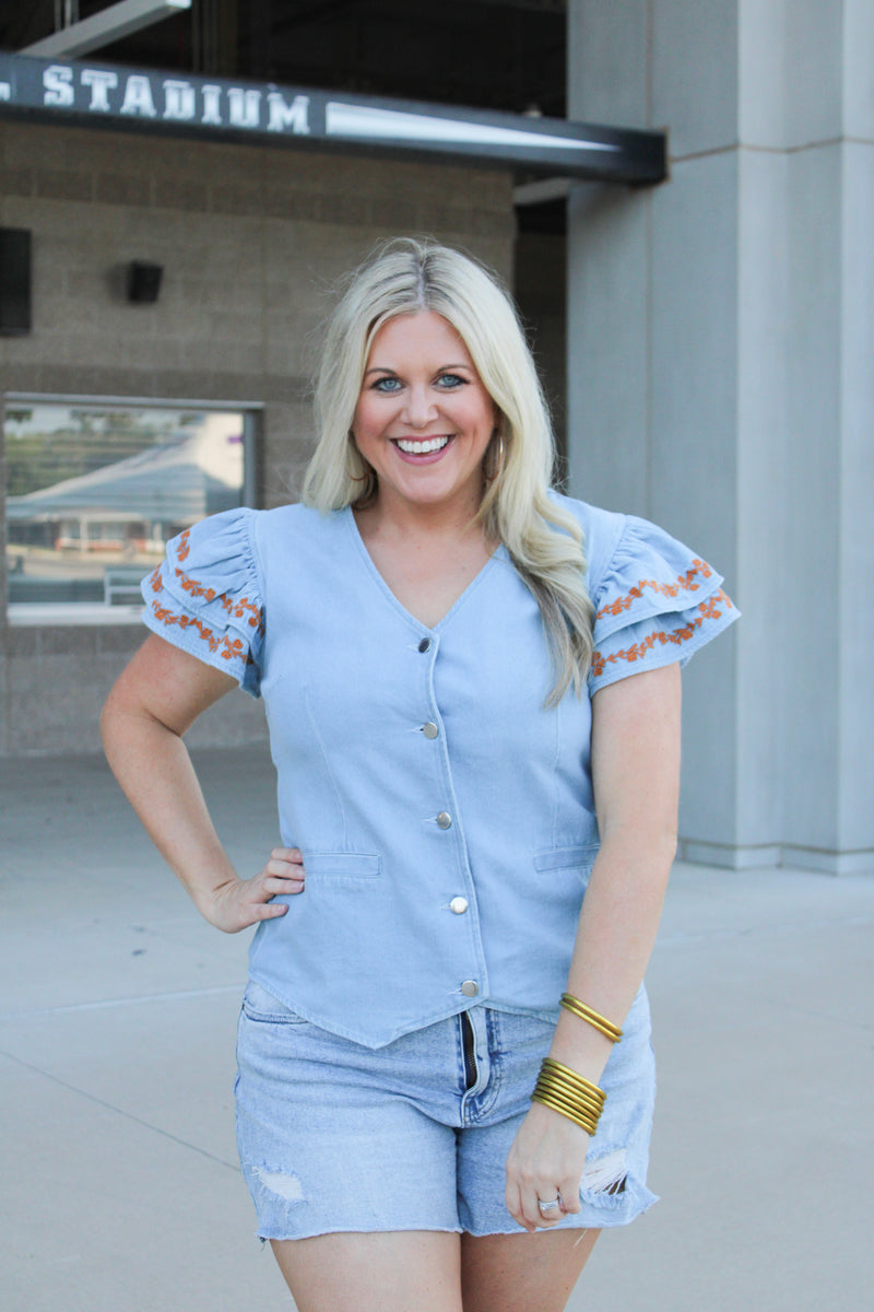 Woman with blonde hair wearing a denim top with burnt orange embroidery on sleeves, and light-wash denim shorts.