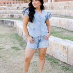 Woman with dark hair wearing a denim top with burnt orange embroidery on sleeves, light-wash denim shorts and white boots. 