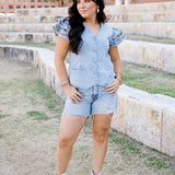 Woman with dark hair wearing a denim top with burnt orange embroidery on sleeves, light-wash denim shorts and white boots. 