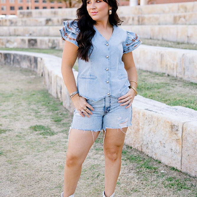 Woman with dark hair wearing a denim top with burnt orange embroidery on sleeves, light-wash denim shorts and white boots. 