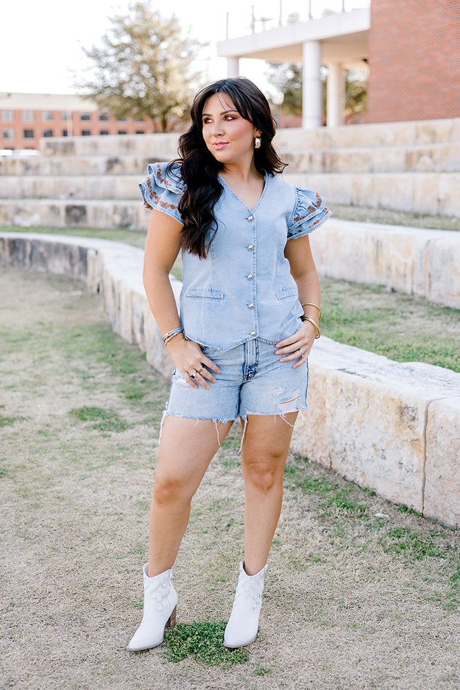 Woman with dark hair wearing a denim top with burnt orange embroidery on sleeves, light-wash denim shorts and white boots. 