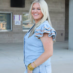 Woman with blonde hair wearing a denim top with burnt orange embroidery on sleeves, and light-wash denim shorts.