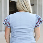 Woman with blonde hair wearing a short-sleeve denim top with maroon floral embroidery on sleeves, and light-wash denim shorts. 