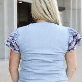 Woman with blonde hair wearing a short-sleeve denim top with maroon floral embroidery on sleeves, and light-wash denim shorts. 