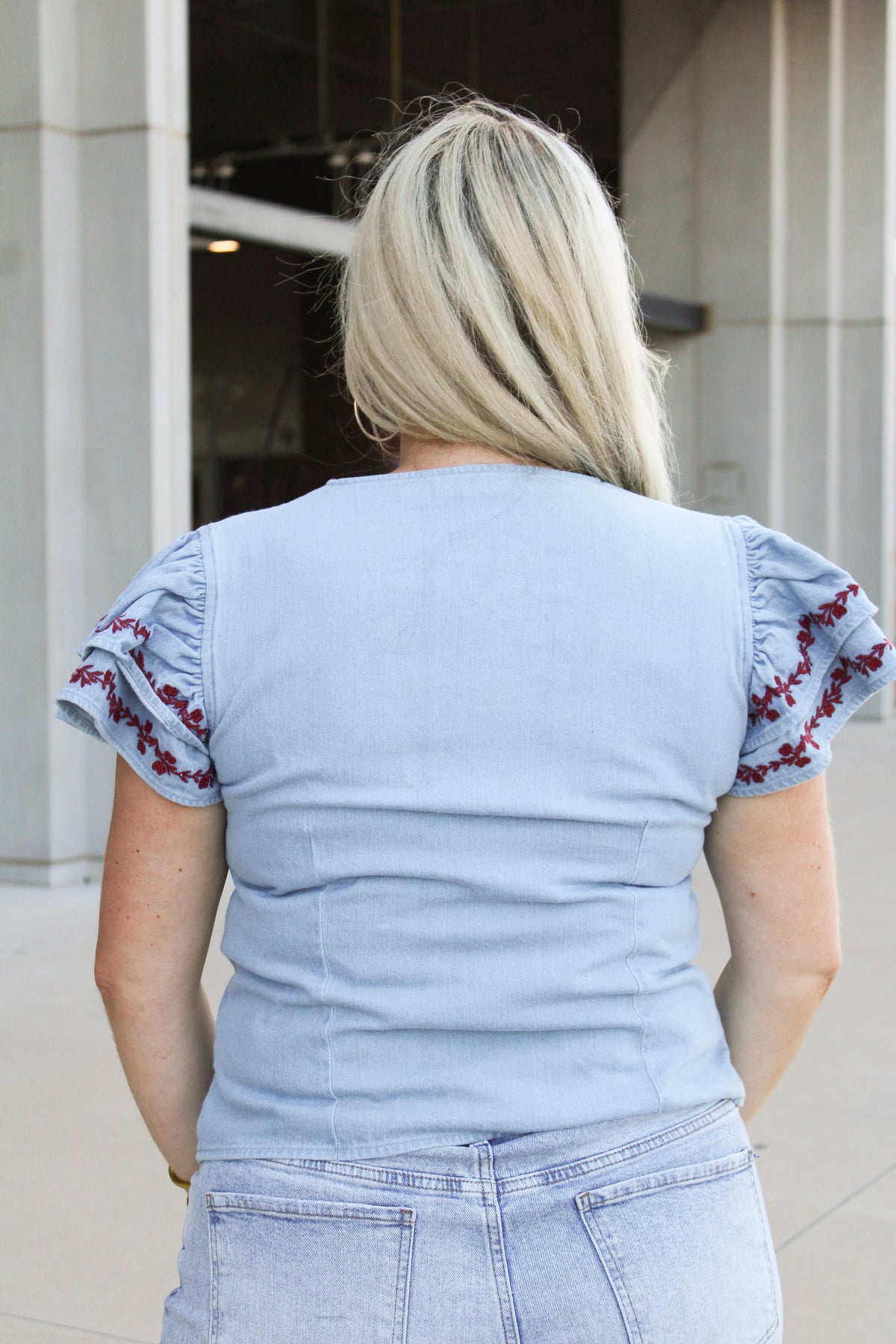 Woman with blonde hair wearing a short-sleeve denim top with maroon floral embroidery on sleeves, and light-wash denim shorts. 