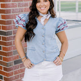 Woman with dark hair wearing a short-sleeve denim top with maroon floral embroidery on sleeves, and light-wash denim shorts. 