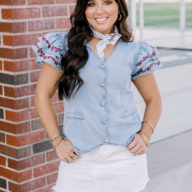 Woman with dark hair wearing a short-sleeve denim top with maroon floral embroidery on sleeves, and light-wash denim shorts. 