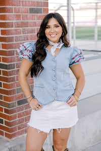Woman with dark hair wearing a short-sleeve denim top with maroon floral embroidery on sleeves, and light-wash denim shorts. 
