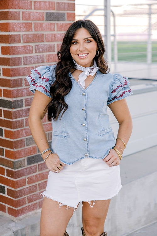 Woman with dark hair wearing a short-sleeve denim top with maroon floral embroidery on sleeves, and light-wash denim shorts. 