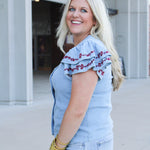 Woman with blonde hair wearing a short-sleeve denim top with maroon floral embroidery on sleeves, and light-wash denim shorts. 