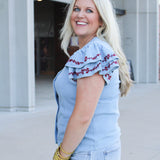 Woman with blonde hair wearing a short-sleeve denim top with maroon floral embroidery on sleeves, and light-wash denim shorts. 