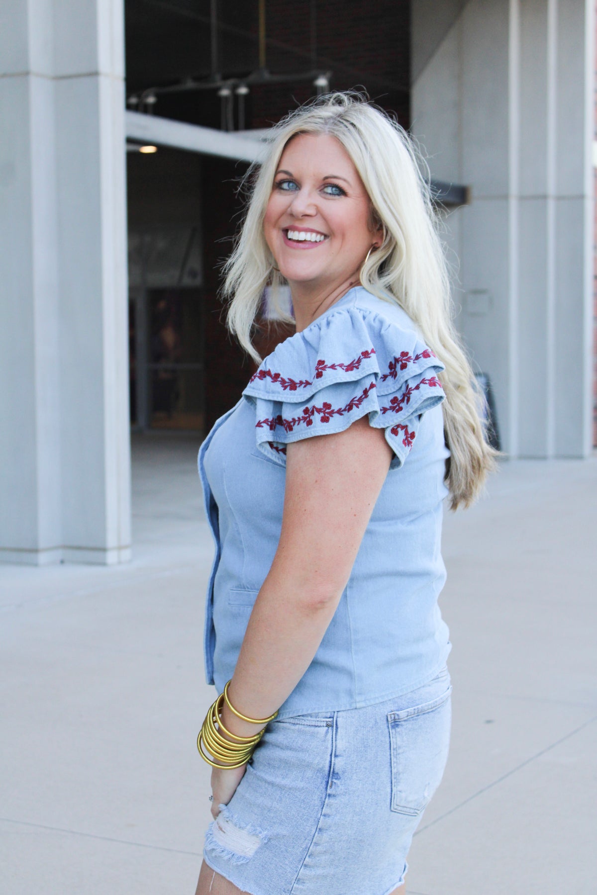 Woman with blonde hair wearing a short-sleeve denim top with maroon floral embroidery on sleeves, and light-wash denim shorts. 