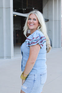 Woman with blonde hair wearing a short-sleeve denim top with maroon floral embroidery on sleeves, and light-wash denim shorts. 