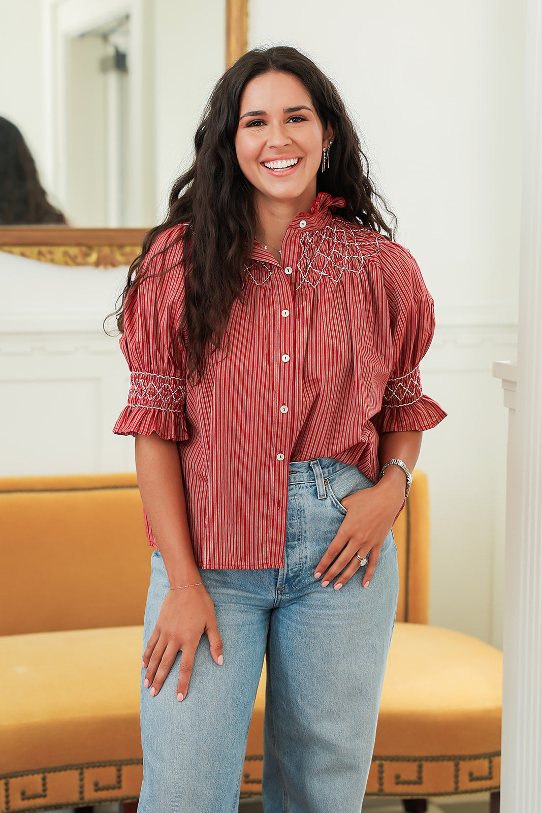 Woman with dark hair standing in a front profile position wearing the Kate Top in a lifestyle scene. The focal point of this image is the embroidery on the front as well as sleeves with with ruffles. The model paired this top with light-wash denim jeans. 