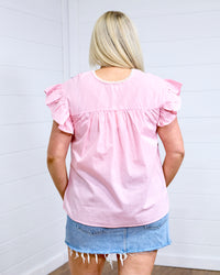 Woman with blonde hair standing in a back profile position wearing the Kinley Top against a white background. The focal point of this image is the gingham pattern on top as well as pleating and ruffle sleeves. The model paired this top with a light-wash denim skirt. 
