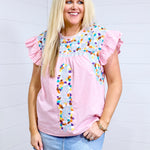 Woman with blonde hair standing in a front profile position wearing the Kinley Top against a white background. The focal point of this image is the embroidery on the front of the top as well as the gingham fabric and ruffle sleeves. The model paired this top with a light-wash denim skirt. 