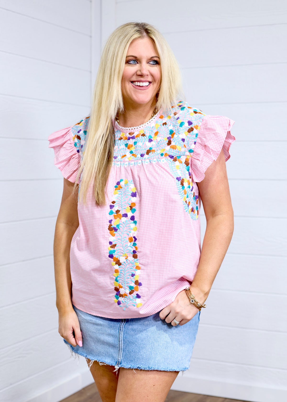 Woman with blonde hair standing in a front profile position wearing the Kinley Top against a white background. The focal point of this image is the embroidery on the front of the top as well as the gingham fabric and ruffle sleeves. The model paired this top with a light-wash denim skirt. 