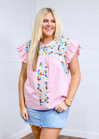 Woman with blonde hair standing in a front profile position wearing the Kinley Top against a white background. The focal point of this image is the embroidery on the front of the top as well as the gingham fabric and ruffle sleeves. The model paired this top with a light-wash denim skirt. 