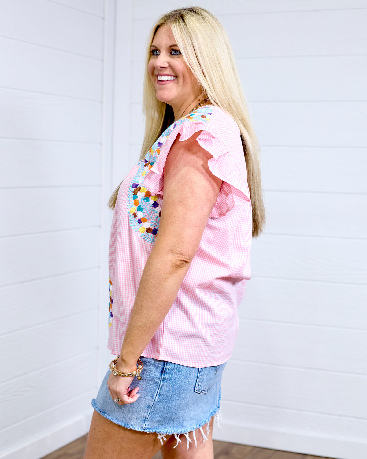 Woman with blonde hair standing in a side profile position wearing the Kinley Top against a white background. The focal point of this image is the embroidery on front of top and ruffle sleeve. The model paired this top with the light-wash denim skirt. 