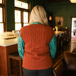 Woman with blonde hair standing in a back profile position wearing the Kodi Vest in a lifestyle scene. The focal point of this image is the cable knit pattern on back of vest. The model paired this top with dark wash denim jeans. 