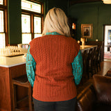 Woman with blonde hair standing in a back profile position wearing the Kodi Vest in a lifestyle scene. The focal point of this image is the cable knit pattern on back of vest. The model paired this top with dark wash denim jeans. 