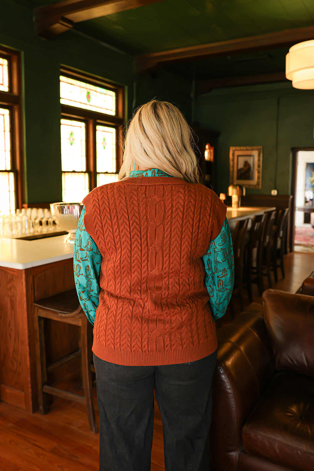 Woman with blonde hair standing in a back profile position wearing the Kodi Vest in a lifestyle scene. The focal point of this image is the cable knit pattern on back of vest. The model paired this top with dark wash denim jeans. 