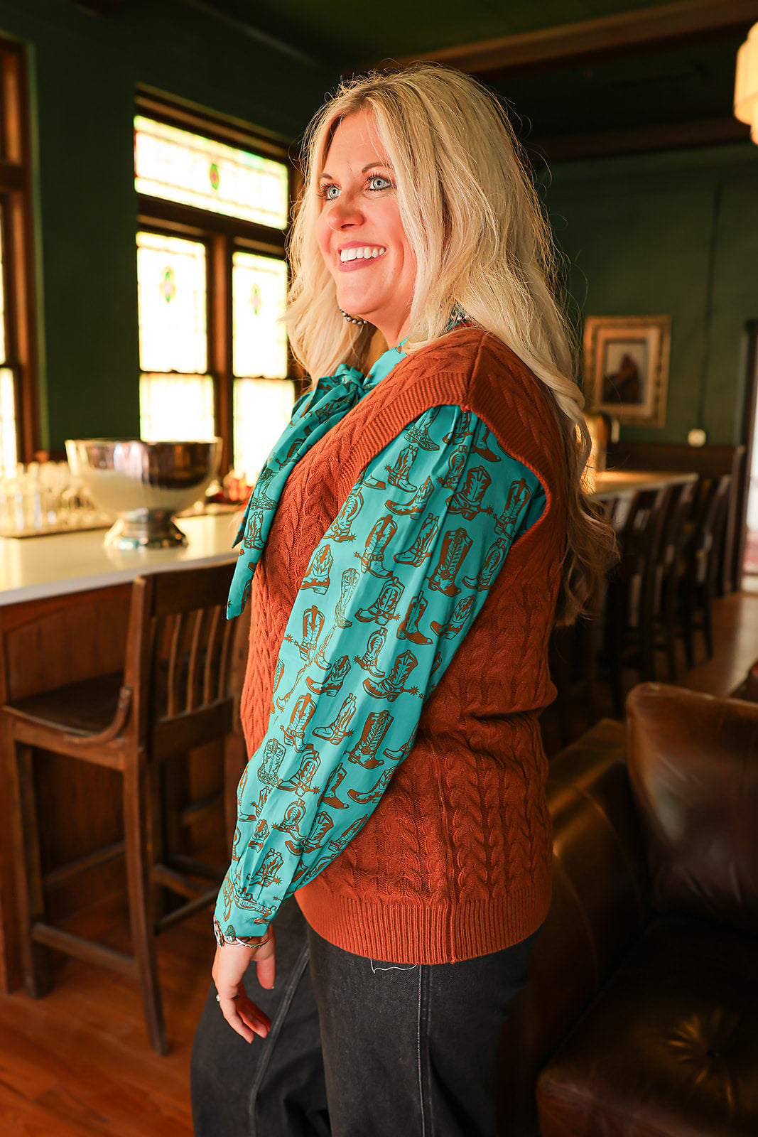 Woman with blonde hair standing in a side profile position wearing the Kodi Vest in a lifestyle scene. The focal point of this image is the sleeveless top and cable knit pattern. The model paired this top with dark-wash denim jeans. 