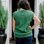 Woman with dark hair wearing a green sleeveless cable-knit vest with "JOY" embroidered on front. Model paired this top with dark-wash denim jeans. 