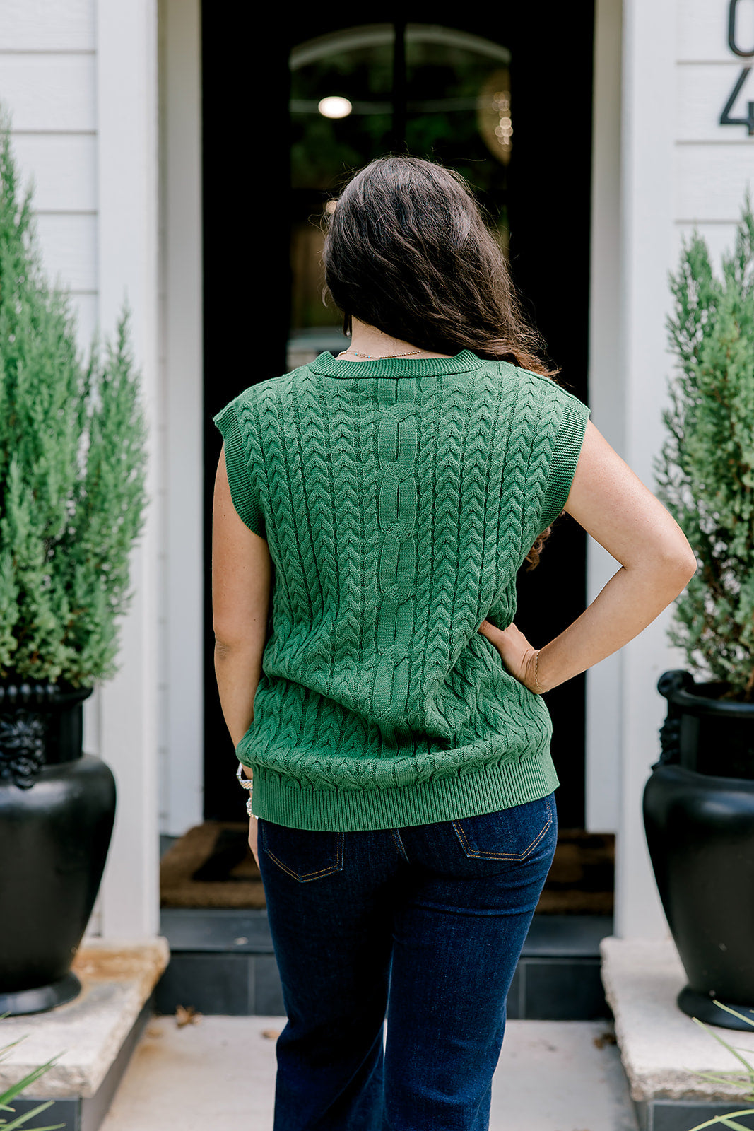 Woman with dark hair wearing a green sleeveless cable-knit vest with "JOY" embroidered on front. Model paired this top with dark-wash denim jeans. 