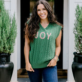 Woman with dark hair wearing a green sleeveless cable-knit vest with "JOY" embroidered on front. Model paired this top with dark-wash denim jeans. 