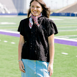Woman with dark hair facing forward wearing a short-sleeve black top with eyelet pattern, and a light-wash denim skirt. 