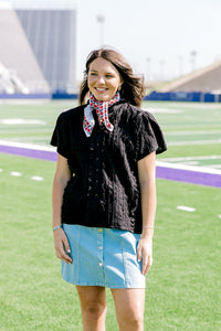 Woman with dark hair facing forward wearing a short-sleeve black top with eyelet pattern, and a light-wash denim skirt. 