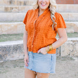 Woman with blonde hair standing in a front profile position wearing a short-sleeve burnt orange eyelet top and a light-wash denim skirt.