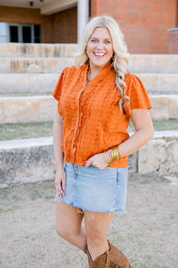 Woman with blonde hair standing in a front profile position wearing a short-sleeve burnt orange eyelet top and a light-wash denim skirt.
