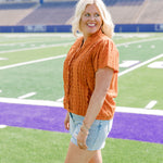 Woman with blonde hair standing in a side profile position wearing a short-sleeve eyelet top in burnt orange. The model paired this top with light-wash denim shorts. 
