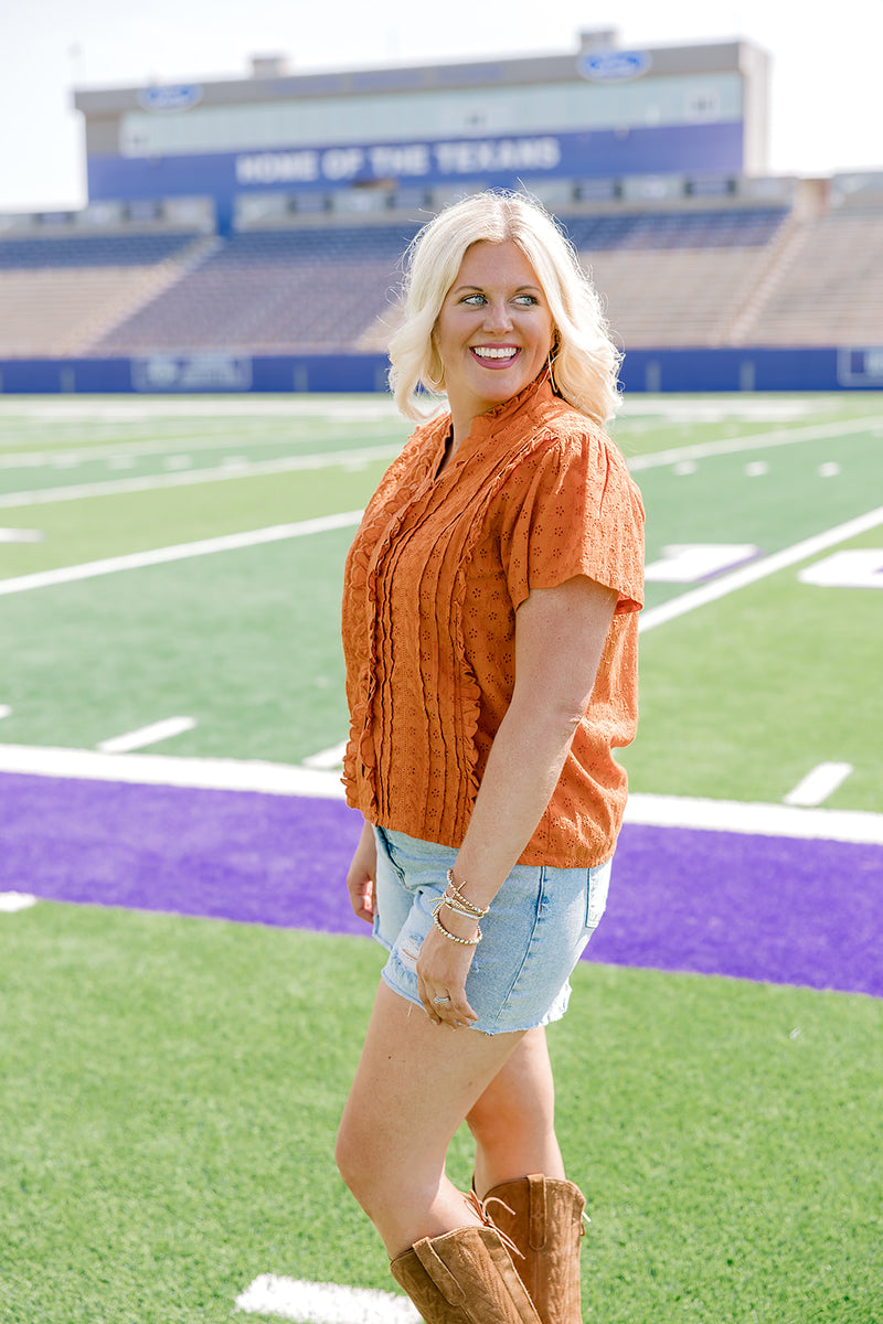 Woman with blonde hair standing in a side profile position wearing a short-sleeve eyelet top in burnt orange. The model paired this top with light-wash denim shorts. 