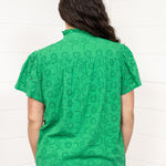 Woman with dark hair standing in a back profile position wearing the Lilly Top against a white background. The focal point of this image is the intricate fabric details and green color. The model paired this top with dark-wash denim jeans. 
