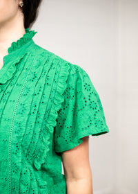 Woman with dark hair standing in front profile position wearing the Lilly Top against a white background. The focal point of this image is a close-up on the green fabric detail.