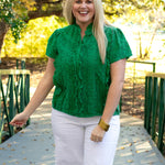 Woman with blonde hair standing in a front profile position wearing the Lilly Top in a lifestyle scene. The focal point of this image is the ruffles down the sides of the buttons on front of shirt, as well as the ruffled high-collar. The model paired this top with white pants.
