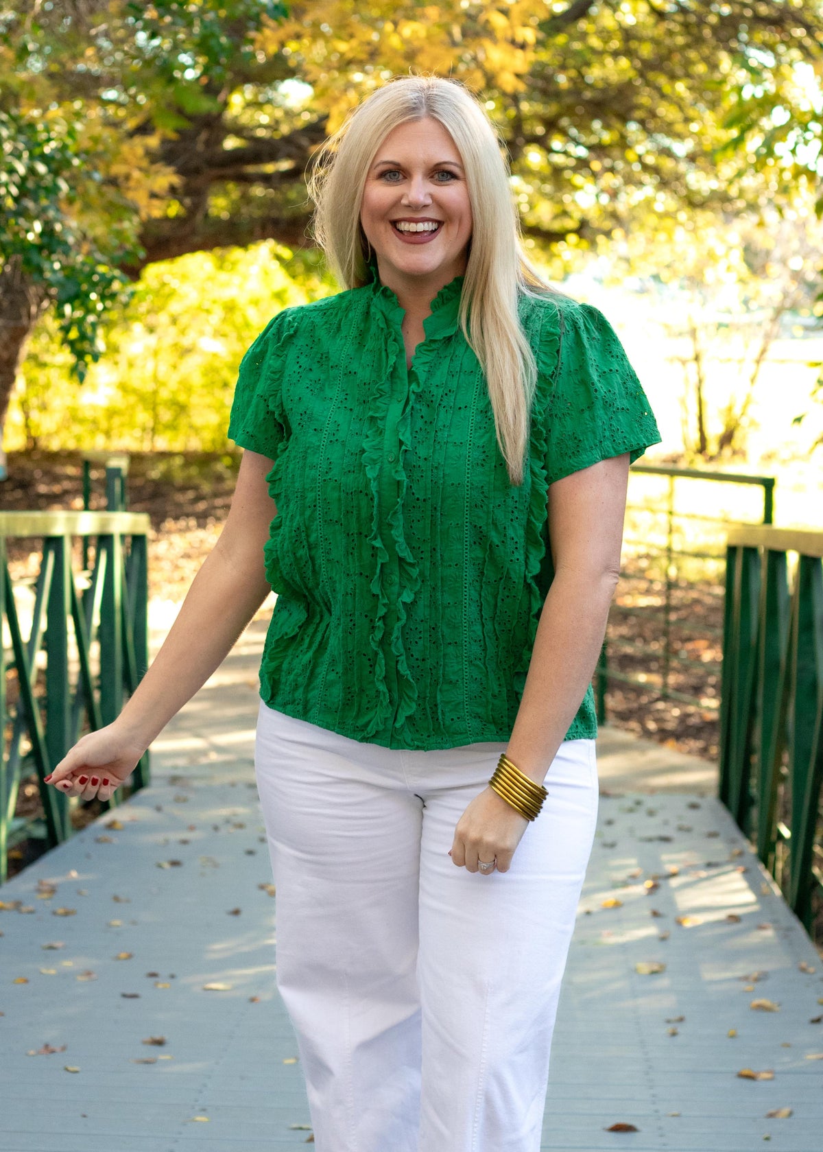 Woman with blonde hair standing in a front profile position wearing the Lilly Top in a lifestyle scene. The focal point of this image is the ruffles down the sides of the buttons on front of shirt, as well as the ruffled high-collar. The model paired this top with white pants.