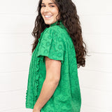Woman with dark hair standing in a side profile position wearing the Lilly Top against a white background. The focal point of this image is the shoulder length and sneak peek of ruffles on the front of top. The model paired this top with dark-wash denim jeans. 